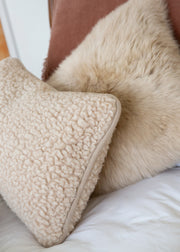 A close-up of a cream sherpa textured throw pillow resting on a white bed next to a fluffy beige sheepskin pillow and a brown headboard.