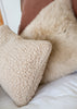 A close-up of a cream sherpa textured throw pillow resting on a white bed next to a fluffy beige sheepskin pillow and a brown headboard.