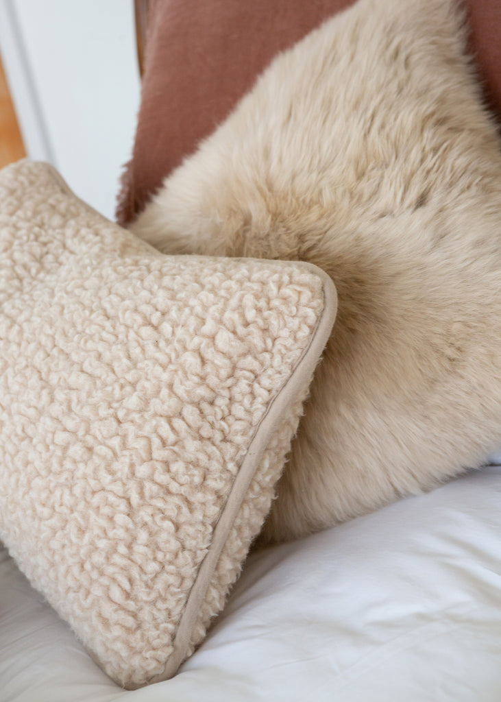 A close-up of a cream sherpa textured throw pillow resting on a white bed next to a fluffy beige sheepskin pillow and a brown headboard.