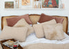 A styled bed with multiple textured throw pillows in beige, cream, and dusty rose, set against a wooden cane and wood headboard. A matching textured blanket is draped at the foot of the bed, with a tray of coffee and a newspaper in the foreground and a white shelf with framed art in the background.