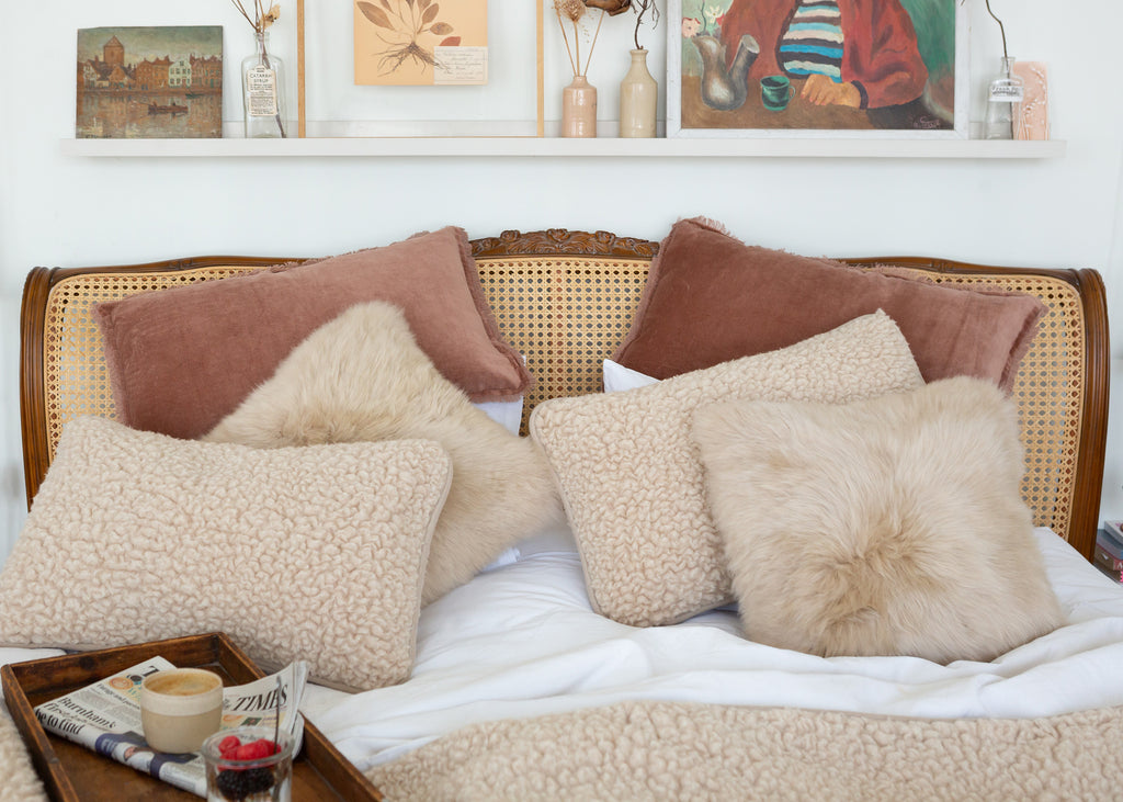A styled bed with multiple textured throw pillows in beige, cream, and dusty rose, set against a wooden cane and wood headboard. A matching textured blanket is draped at the foot of the bed, with a tray of coffee and a newspaper in the foreground and a white shelf with framed art in the background.