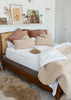 A styled bedroom scene featuring a wooden bed with a cane headboard, dressed in white bedding, blush pink pillows, and beige sherpa throw and rug accents.