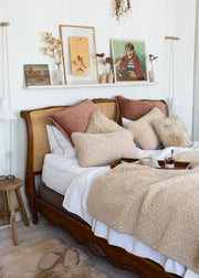 A cozy bedroom scene featuring a wooden bed with a cane headboard, styled with white bedding, textured beige throw pillows, and a cream bouclé blanket. A wooden side stool and a small shelf with framed art and dried flowers are visible against a white wall.