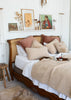 A cozy bedroom scene featuring a wooden bed with a cane headboard, styled with white bedding, textured beige throw pillows, and a cream bouclé blanket. A wooden side stool and a small shelf with framed art and dried flowers are visible against a white wall.
