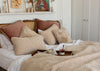 A cozy bedroom scene with a bed styled in neutral tones, featuring textured cream sherpa pillows, dusty rose and brown pillows, white bedding, a beige blanket, and a wooden tray on the bed. A white shelf with framed art and dried flowers is mounted on the wall above the bed.