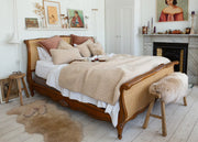 A vintage-style wooden and cane bed frame styled in a bright, modern bedroom with beige sherpa bedding, decorative pillows, a fur bench, and a fireplace in the background.