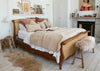 A vintage-style wooden and cane bed frame styled in a bright, modern bedroom with beige sherpa bedding, decorative pillows, a fur bench, and a fireplace in the background.