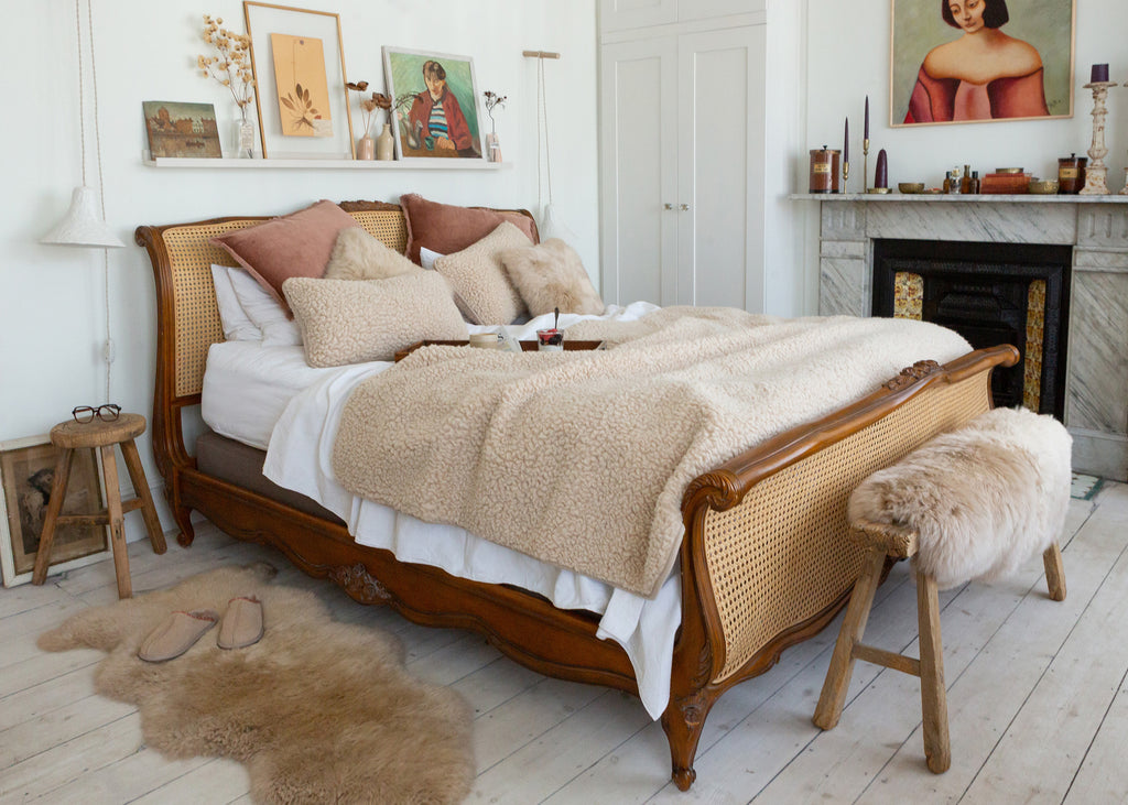A vintage-style wooden and cane bed frame styled in a bright, modern bedroom with beige sherpa bedding, decorative pillows, a fur bench, and a fireplace in the background.