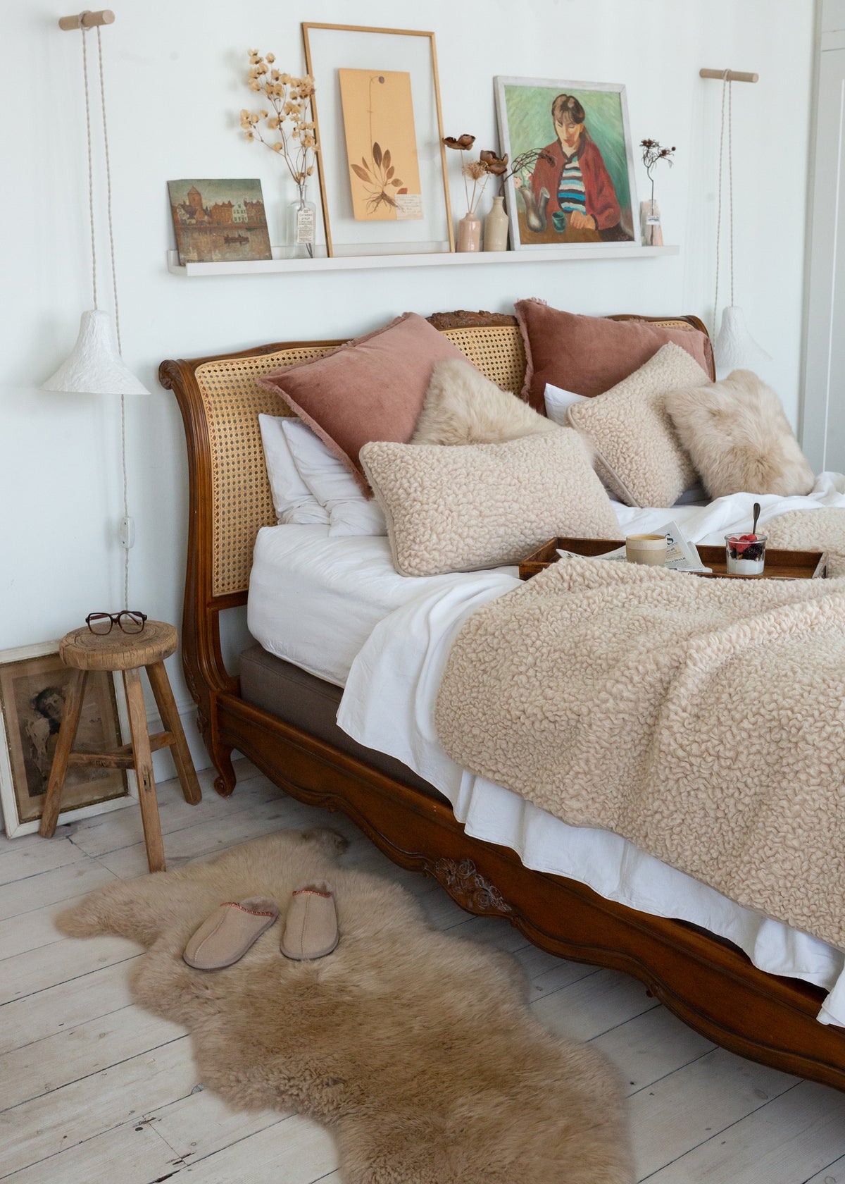 A cozy bedroom scene featuring a wooden bed with a cane headboard, styled with beige textured throw blankets, cream sheepskin pillows, and dusty rose accent pillows. A matching beige sheepskin rug lies on the light wood floor next to a small wooden stool.