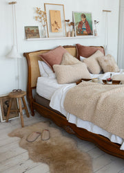 A cozy bedroom scene featuring a wooden bed with a cane headboard, styled with beige textured throw blankets, cream sheepskin pillows, and dusty rose accent pillows. A matching beige sheepskin rug lies on the light wood floor next to a small wooden stool.