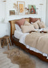 A cozy bedroom scene featuring a wooden bed with a cane headboard, styled with beige textured throw blankets, cream sheepskin pillows, and dusty rose accent pillows. A matching beige sheepskin rug lies on the light wood floor next to a small wooden stool.