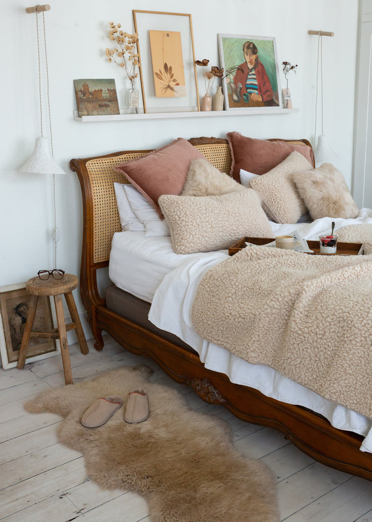 A cozy bedroom scene featuring a wooden bed with a cane headboard, styled with beige textured throw blankets, cream sheepskin pillows, and dusty rose accent pillows. A matching beige sheepskin rug lies on the light wood floor next to a small wooden stool.