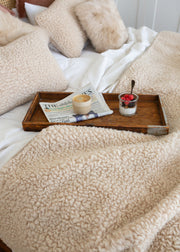 A cozy bedroom scene with cream sherpa bedding, including a textured blanket, pillows, and duvet, styled with a wooden tray holding a newspaper, a small cup, and a yogurt bowl on a bed with white sheets.