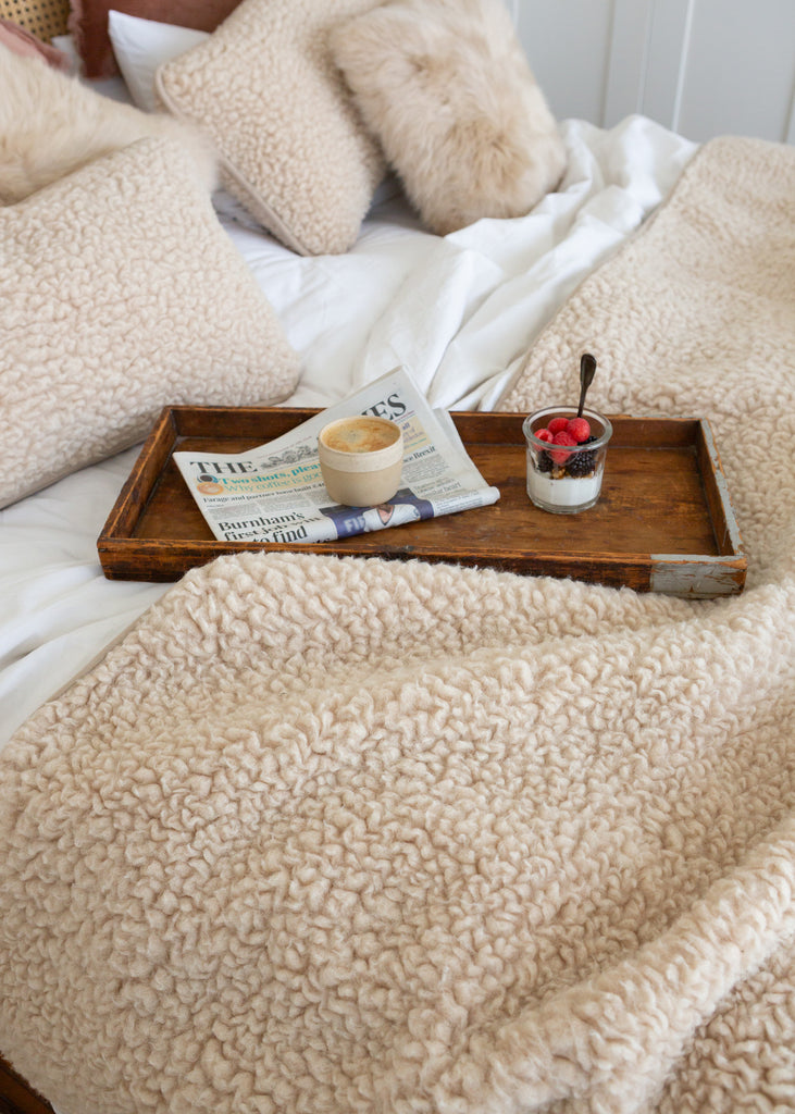 A cozy bedroom scene with cream sherpa bedding, including a textured blanket, pillows, and duvet, styled with a wooden tray holding a newspaper, a small cup, and a yogurt bowl on a bed with white sheets.