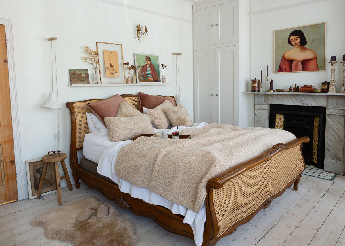 A rustic wooden bed with a woven cane headboard and footboard, dressed in beige and white bedding, styled in a bright, modern bohemian bedroom with a fireplace and wall art.