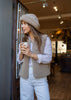 A woman wearing a beige merino gilet, matching merino hat, striped shirt, and white pants stands in a cafe doorway holding a paper coffee cup.