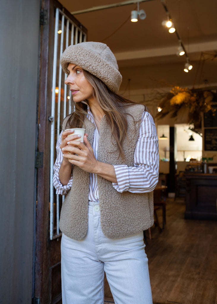 A woman wearing a beige merino gilet, matching merino hat, striped shirt, and white pants stands in a cafe doorway holding a paper coffee cup.