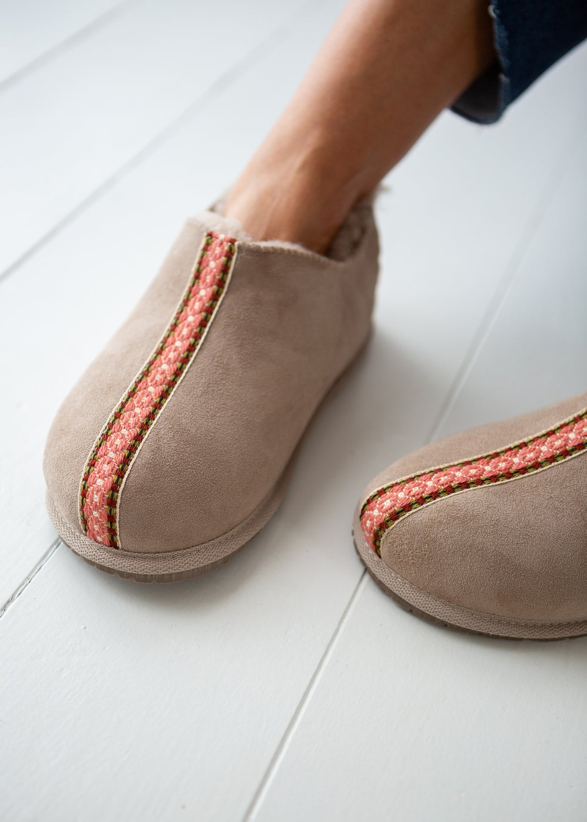 A close-up of a person wearing taupe suede slip-on slippers boots with a pink and white patterned vertical stripe, standing on a white wooden floor.