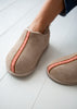 A close-up of a person wearing taupe suede slip-on slippers boots with a pink and white patterned vertical stripe, standing on a white wooden floor.