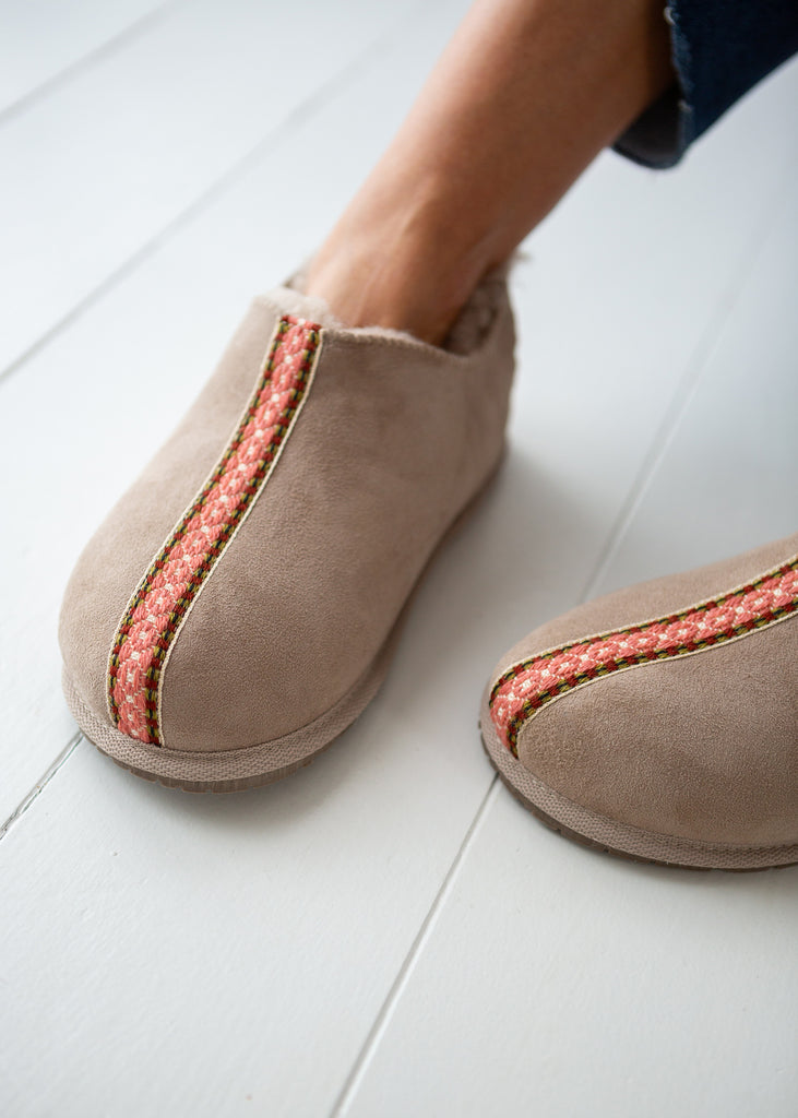 A close-up of a person wearing taupe suede slip-on slippers boots with a pink and white patterned vertical stripe, standing on a white wooden floor.
