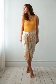 Woman wearing a yellow top and colorful skirt standing in a room with white walls and wooden floor.
