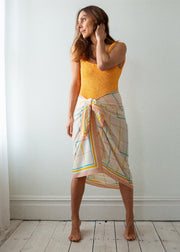 Woman wearing a yellow top and patterned skirt standing against a white wall.