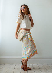 Woman wearing a white top and colorful skirt against a plain white wall.
