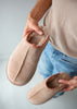 Person holding a pair of beige slippers with brown trim on a neutral background