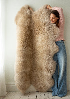 Person holding a large beige sheepskin rug against a white wall.
