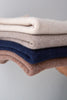 Four folded knit sweaters in cream, beige, navy, and dusty rose, stacked neatly against a neutral gray background.