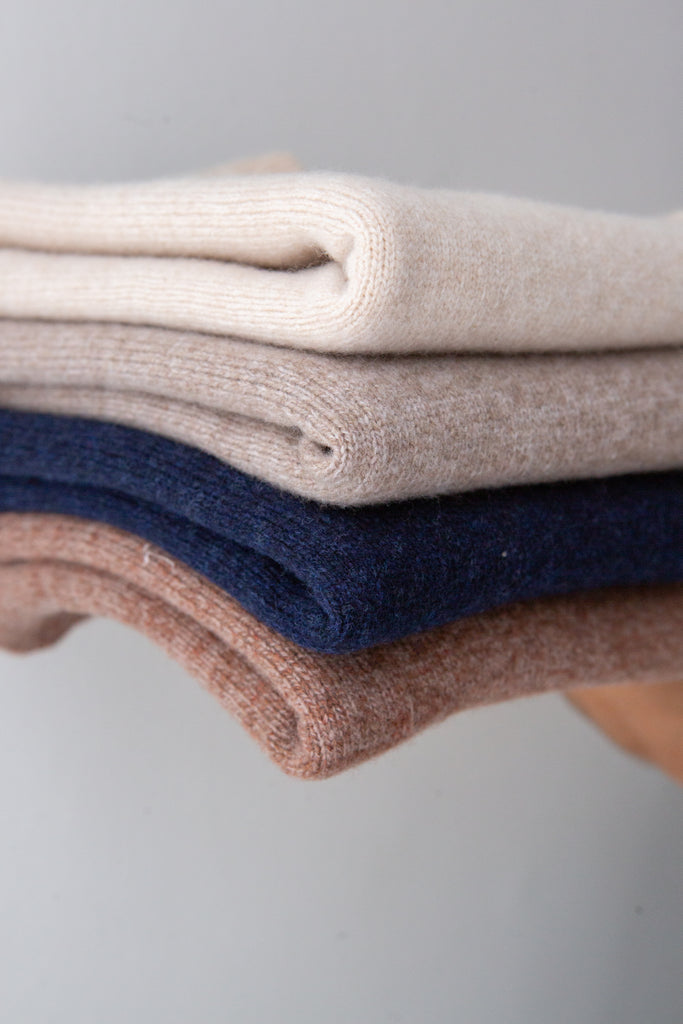 Four folded knit sweaters in cream, beige, navy, and dusty rose, stacked neatly against a neutral gray background.