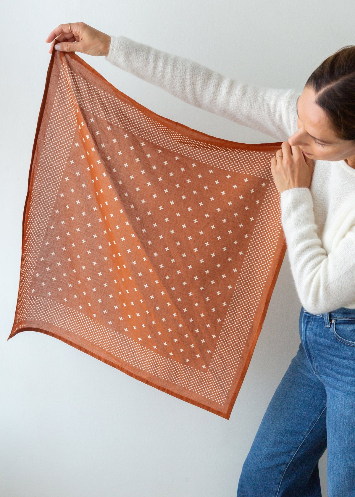 A woman holds up a square scarf against a plain light gray background. The scarf is rust orange with a white polka-dot pattern and solid orange border.