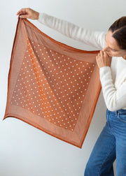 A woman holds up a square scarf against a plain light gray background. The scarf is rust orange with a white polka-dot pattern and solid orange border.