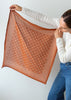 A woman holds up a square scarf against a plain light gray background. The scarf is rust orange with a white polka-dot pattern and solid orange border.