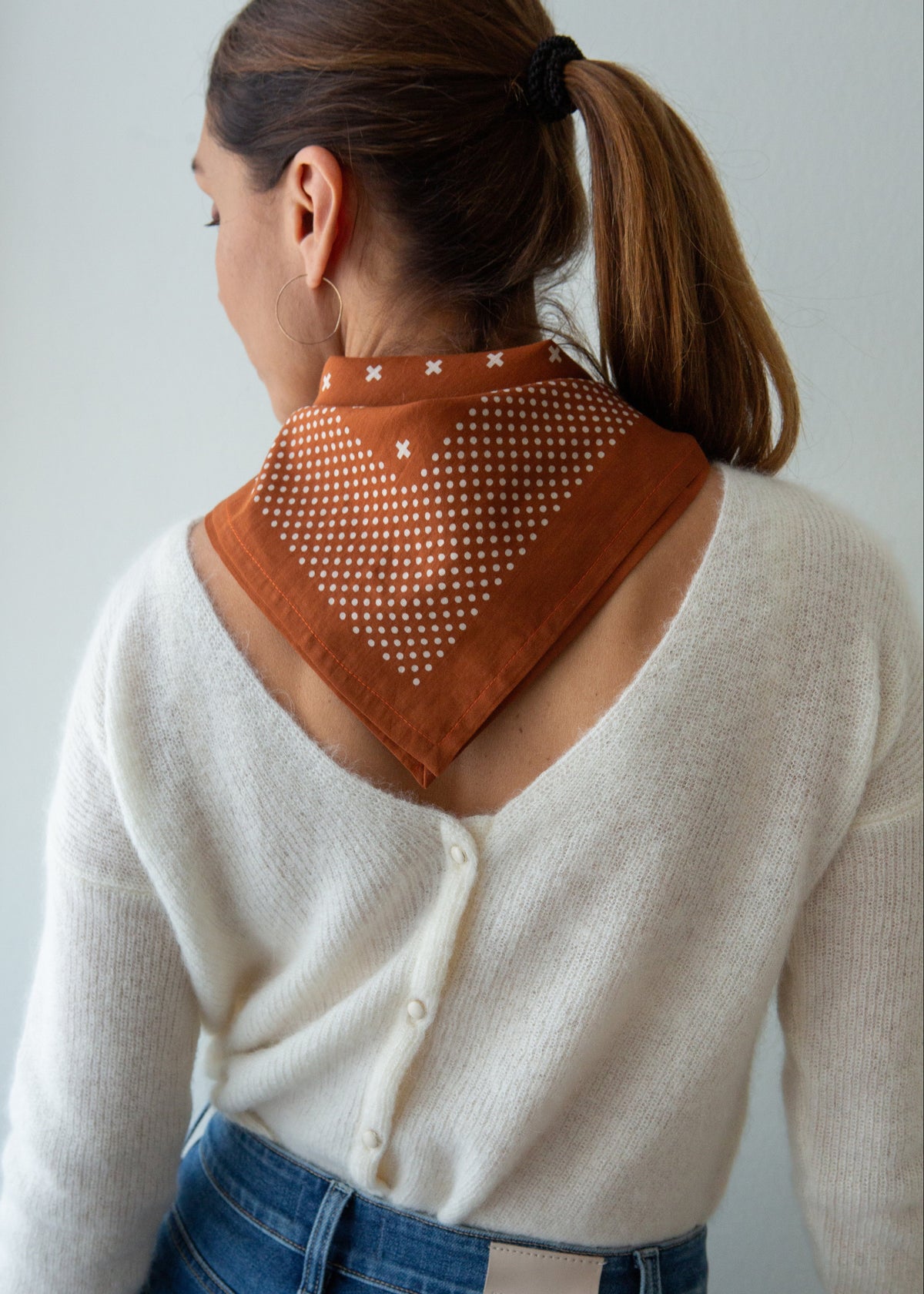 A woman shown from the back wearing a rust-orange bandana with a white geometric pattern around her neck, paired with a white knit cardigan against a light gray background.
