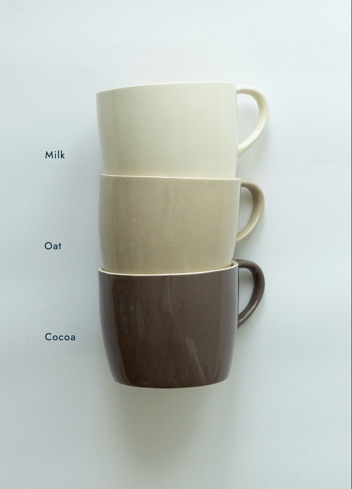 Three stacked ceramic mugs in cream, oatmeal, and dark brown, labeled 'Milk', 'Oat', and 'Coco' on the left side, against a plain white background.