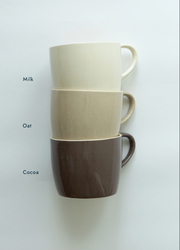 Three stacked ceramic mugs in cream, oatmeal, and dark brown, labeled 'Milk', 'Oat', and 'Coco' on the left side, against a plain white background.