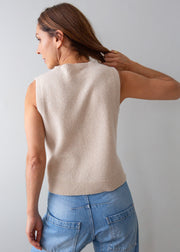 A woman shown from the back wearing a sleeveless knit sweater vest in a light beige color, paired with blue jeans, against a plain light gray background.