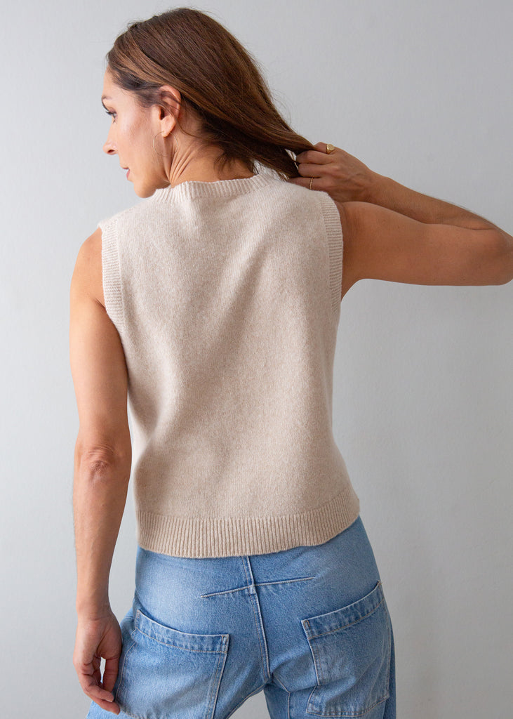 A woman shown from the back wearing a sleeveless knit sweater vest in a light beige color, paired with blue jeans, against a plain light gray background.