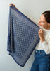 A woman holds up a square navy blue scarf with a white star pattern and dotted borders against a plain light gray wall.