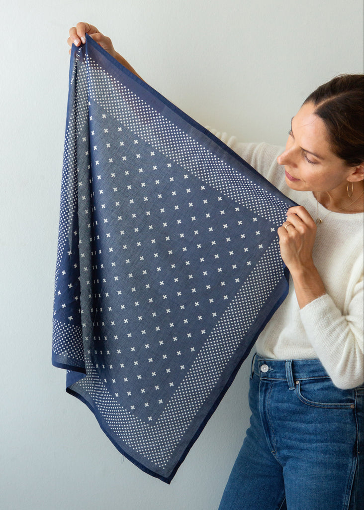 A woman holds up a square navy blue scarf with a white star pattern and dotted borders against a plain light gray wall.