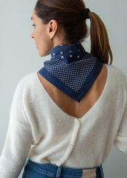 A woman shown from the back wearing a navy blue bandana with white polka dots and small white star accents, styled around her neck over a cream knit cardigan and blue jeans.