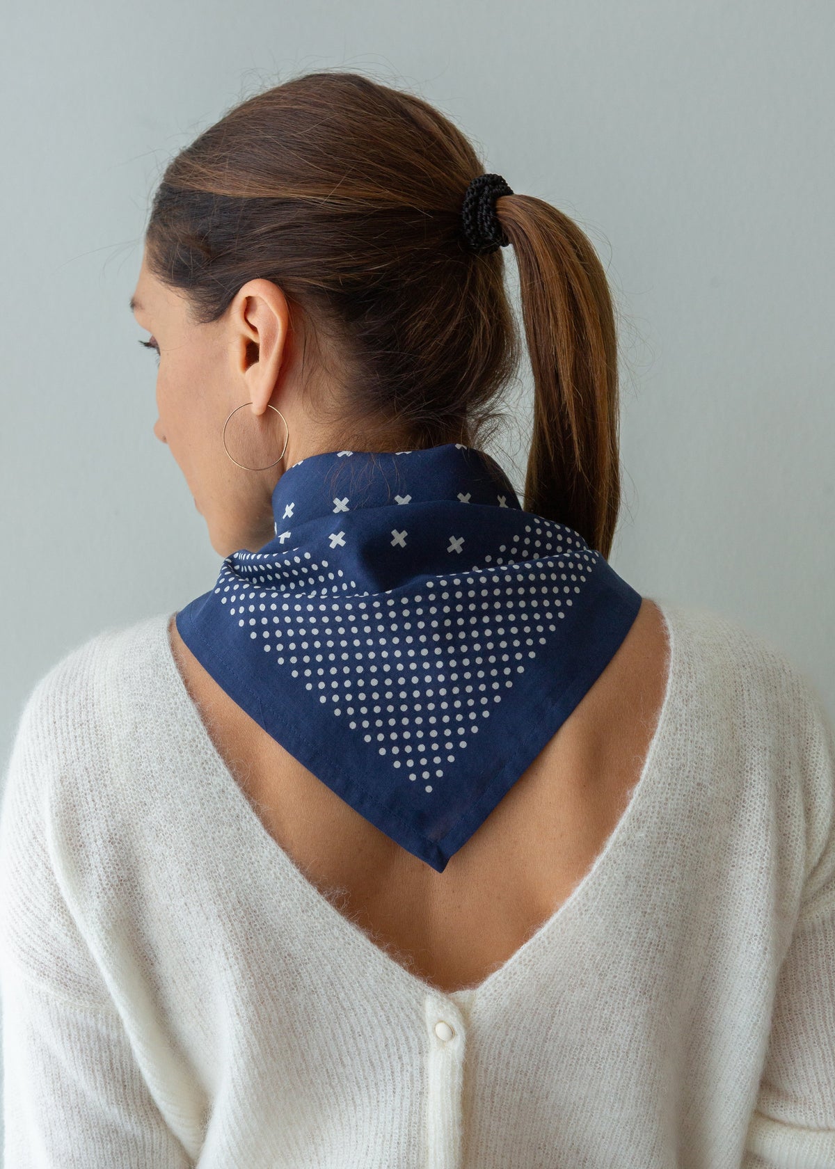 A woman shown from the back wearing a navy blue neck scarf with a white polka dot and star pattern, paired with a cream knit cardigan against a light gray background.