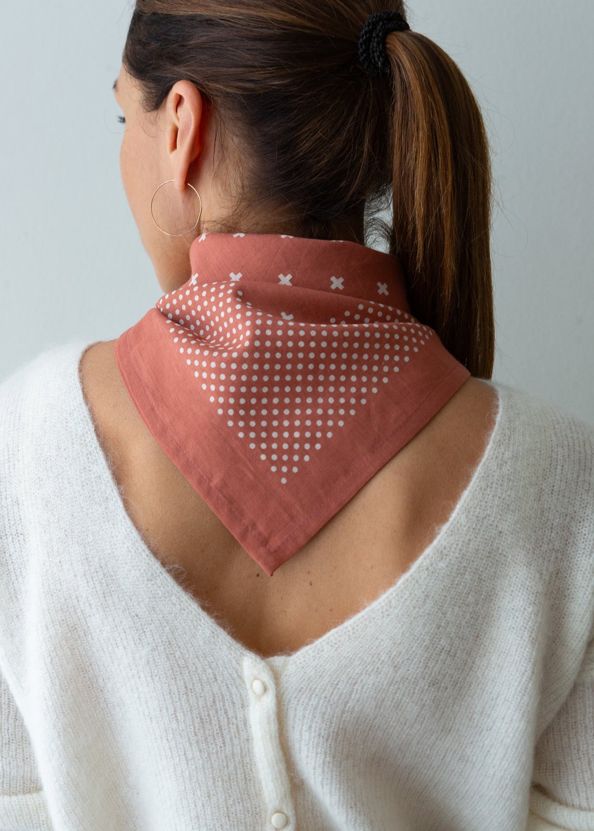 A woman shown from the back wearing a dusty rose bandana with white polka dots and small white cross motifs, styled over her shoulders.