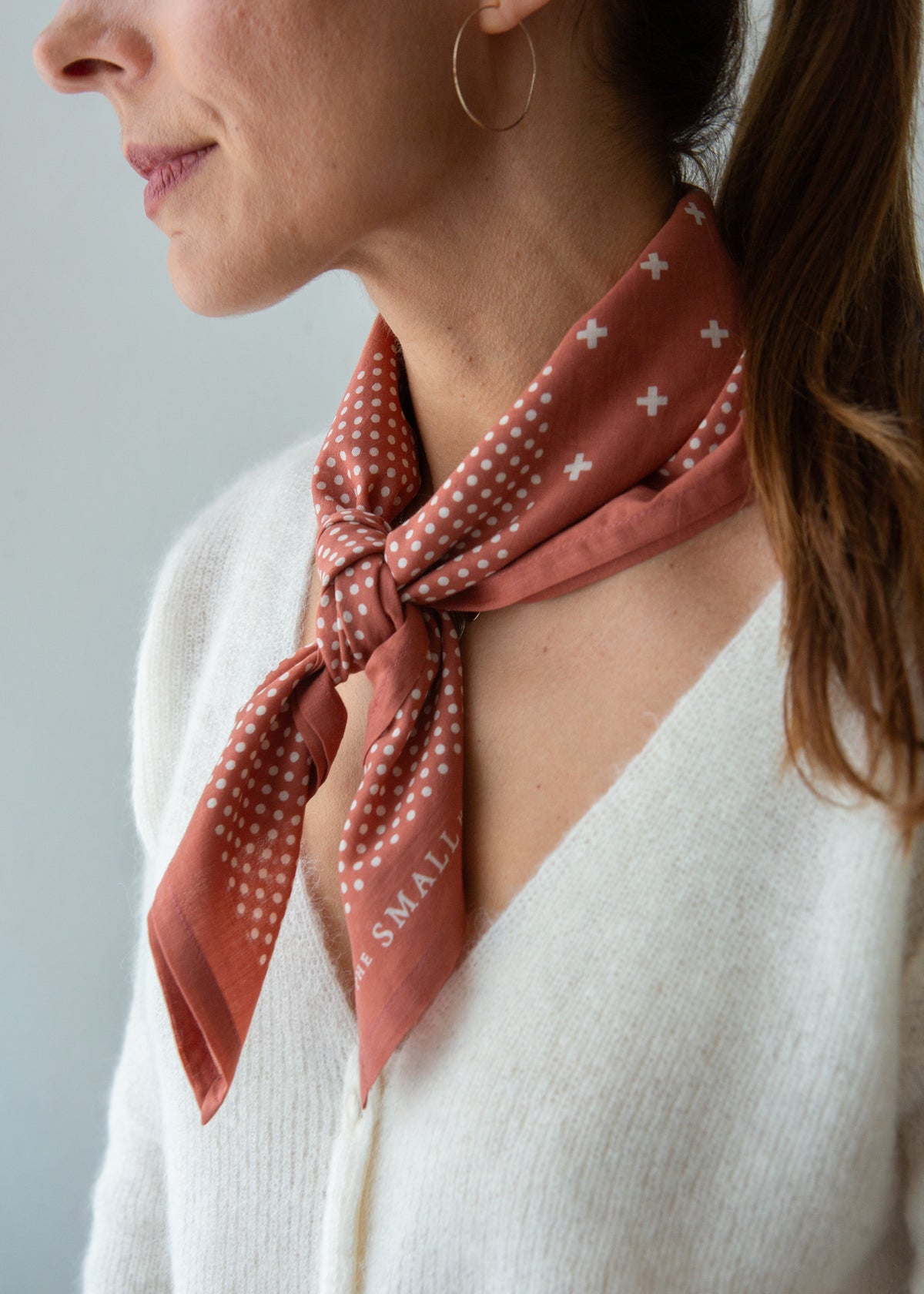 A close-up of a woman wearing a terracotta-colored silk scarf with white polka dots and small white cross motifs, tied around her neck. The scarf features the text 'SMALL' printed in white.