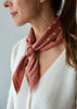 A close-up of a woman wearing a terracotta-colored silk scarf with white polka dots and small white cross motifs, tied around her neck. The scarf features the text 'SMALL' printed in white.