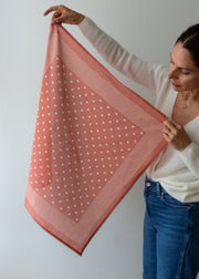 A woman holds up a square scarf with a coral pink and white polka dot pattern against a plain light gray wall.