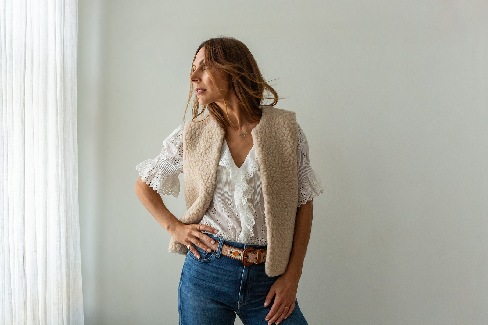 A woman posing in a cream merino gilet over a white ruffled blouse, paired with blue jeans and a brown belt, set against a minimalist light gray wall and white curtain.